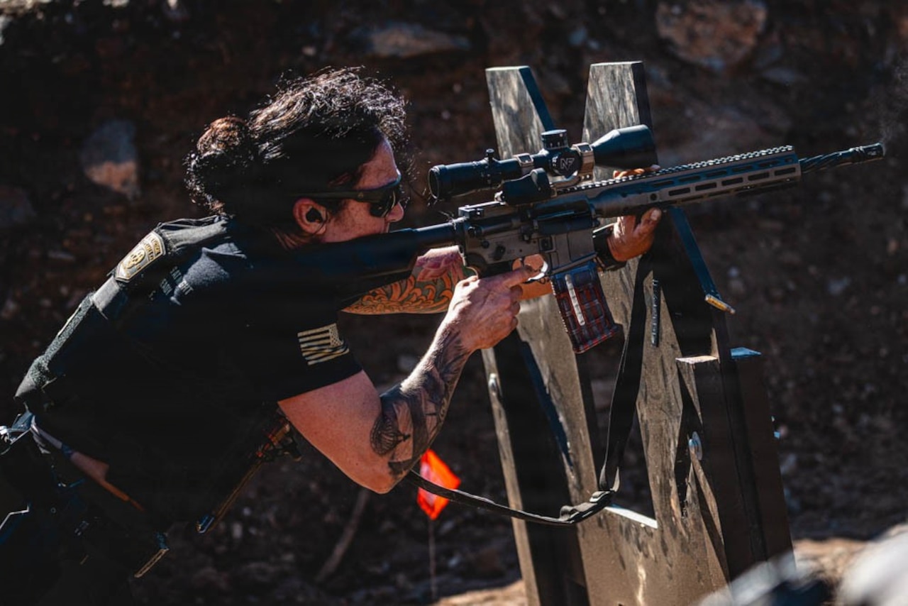 A soldier aims down toward a target using a rifle wearing ear plugs and sunglasses.