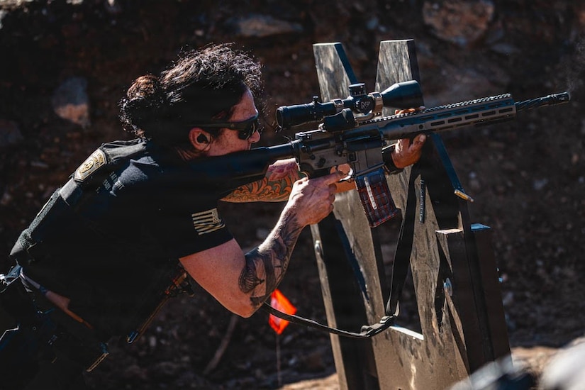 A soldier aims down toward a target using a rifle wearing ear plugs and sunglasses.