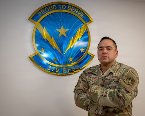 An Airman stands in front of a sign with his arms folded. The sign says "Proud to Serve, 379 EFSS"