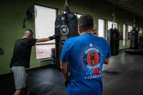 A person punches a punching bag with their right hand while another stands in the foreground watching. His shirt says "Rumble in the Deid Coach"