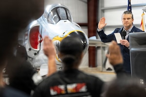 A person in civilian attire holds a microphone and piece of paper in one hand and raises the other hand, with a fighter jet in the background indoors; other people also raising their right hands face the person in the foreground.