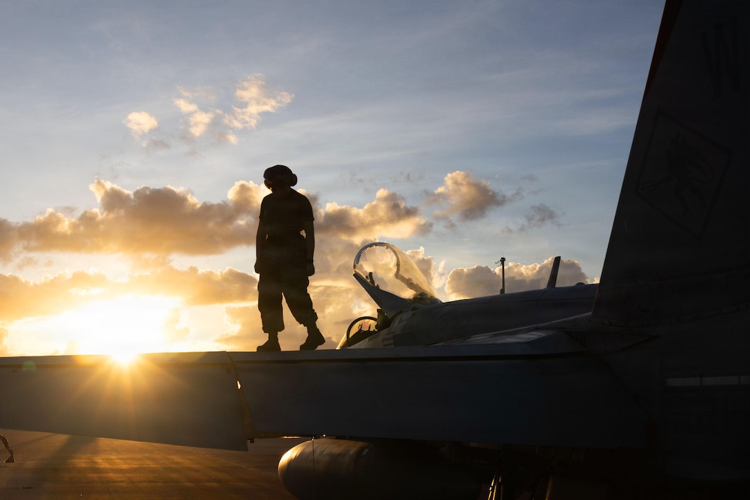A silhouette of a person wearing a helmet while walking on the wing of a plane, while the sun shines brightly in a blue sky with clouds.