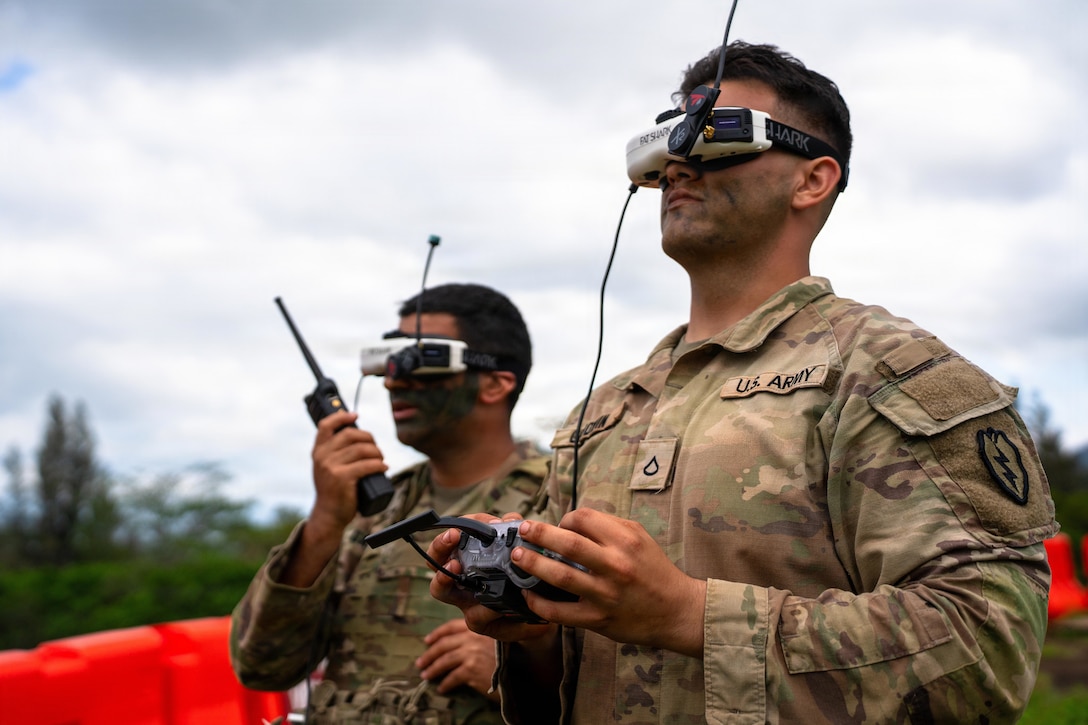 Two men wear camouflage military uniforms and drone head equipment; one is holding the controls for a drone, and the other is holding and speaking through a walkie-talkie.