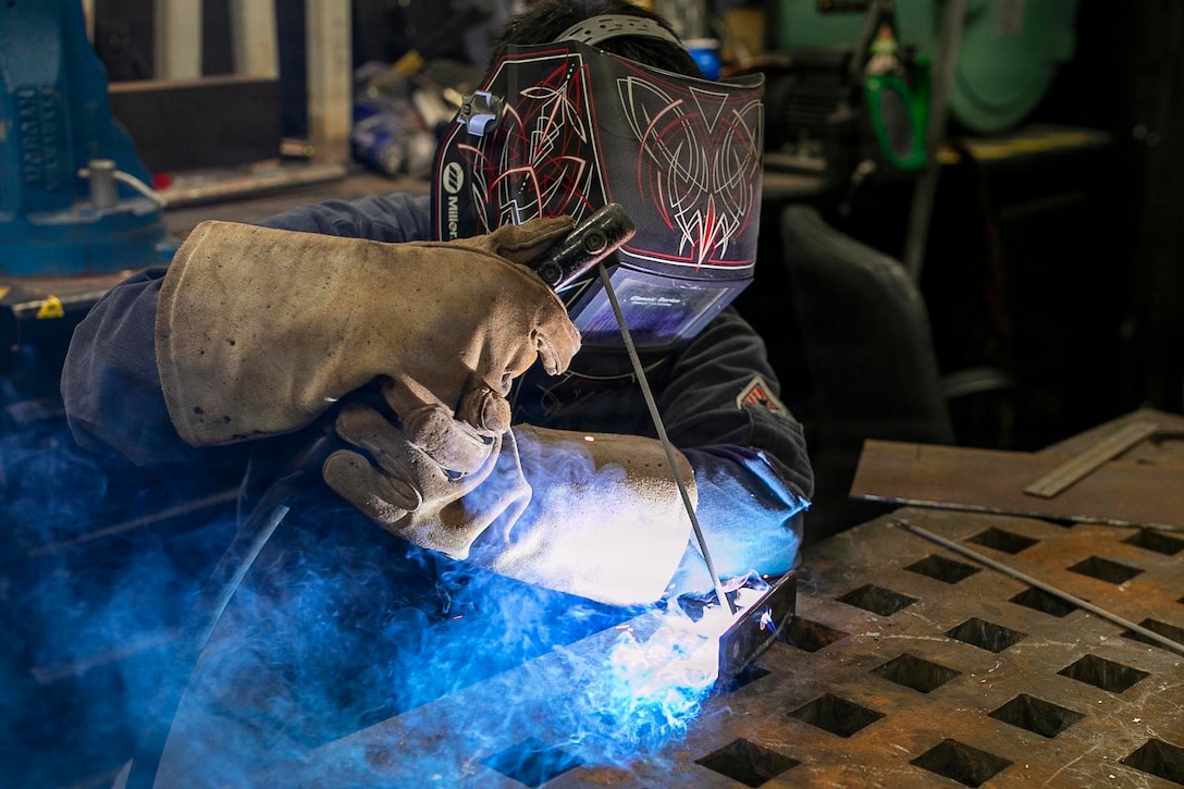 A person wearing a welding helmet and gloves holds a welding tool, as blue and white smoke emits from the point of contact in a repair shop.