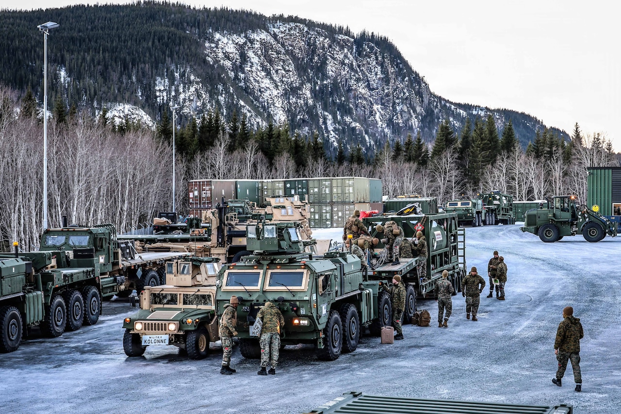 Several people in camouflage military uniforms walk around military vehicles that are lined up on an ice-covered road with a snow-covered mountain in the background.