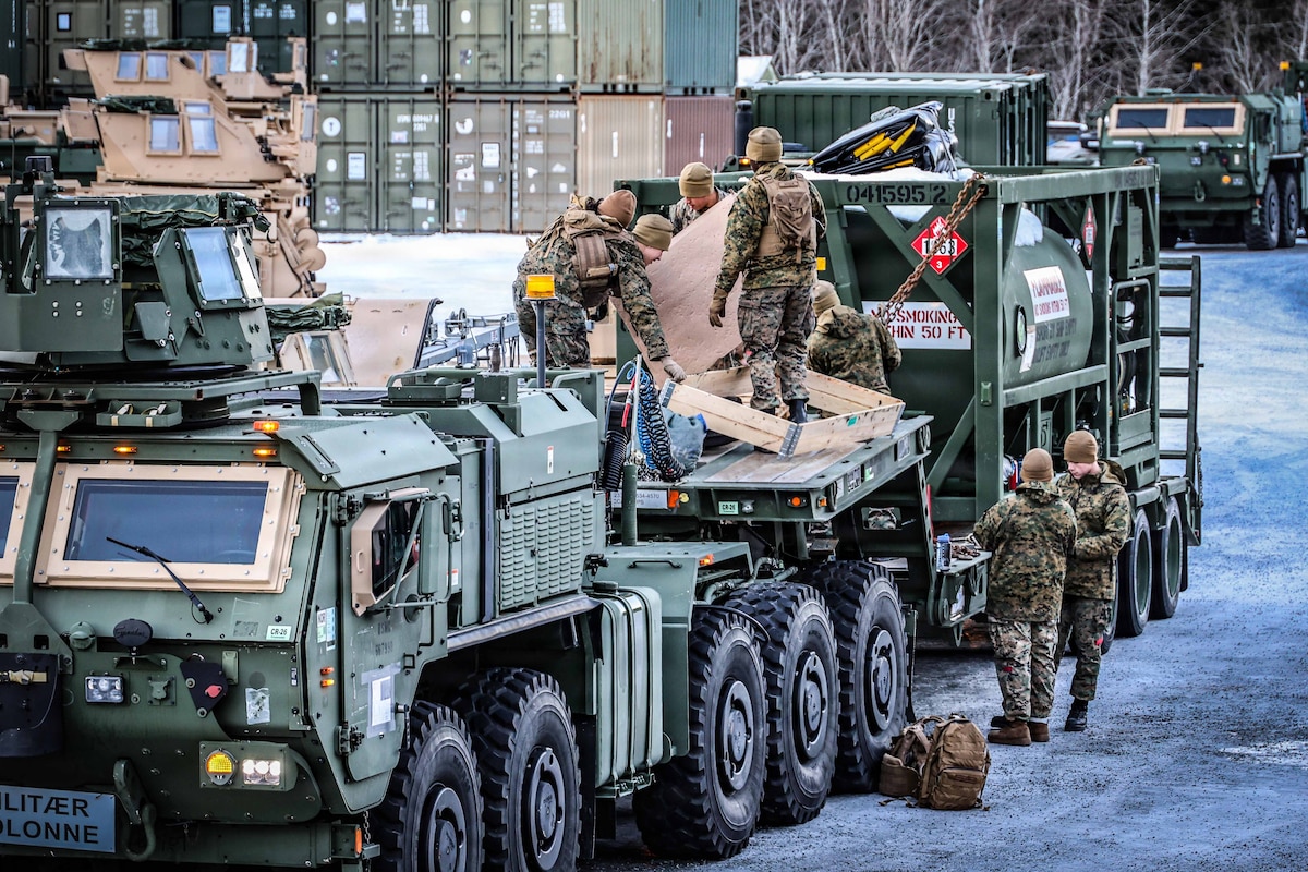 Six people in camouflage military uniforms load equipment onto a military vehicle, outside on an ice-covered road.
