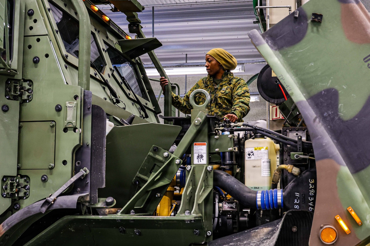 A woman in a camouflage military uniform stands on the ledge of a large military vehicle and looks to her right into the windshield of the vehicle.