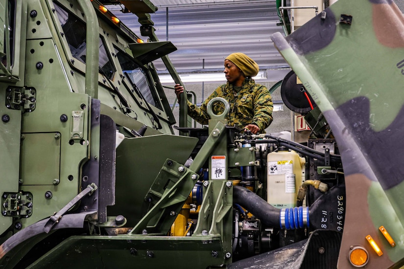 A woman in a camouflage military uniform stands on the ledge of a large military vehicle and looks to her right into the windshield of the vehicle.