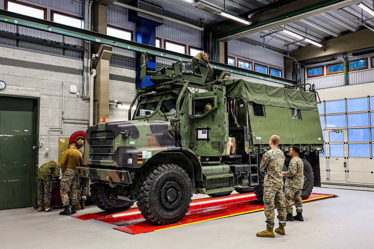 A large military vehicle sits on top of an inspection zone inside a warehouse as several people in camouflage military uniforms walk around it, and another person in similar attire stands on the roof of the vehicle.