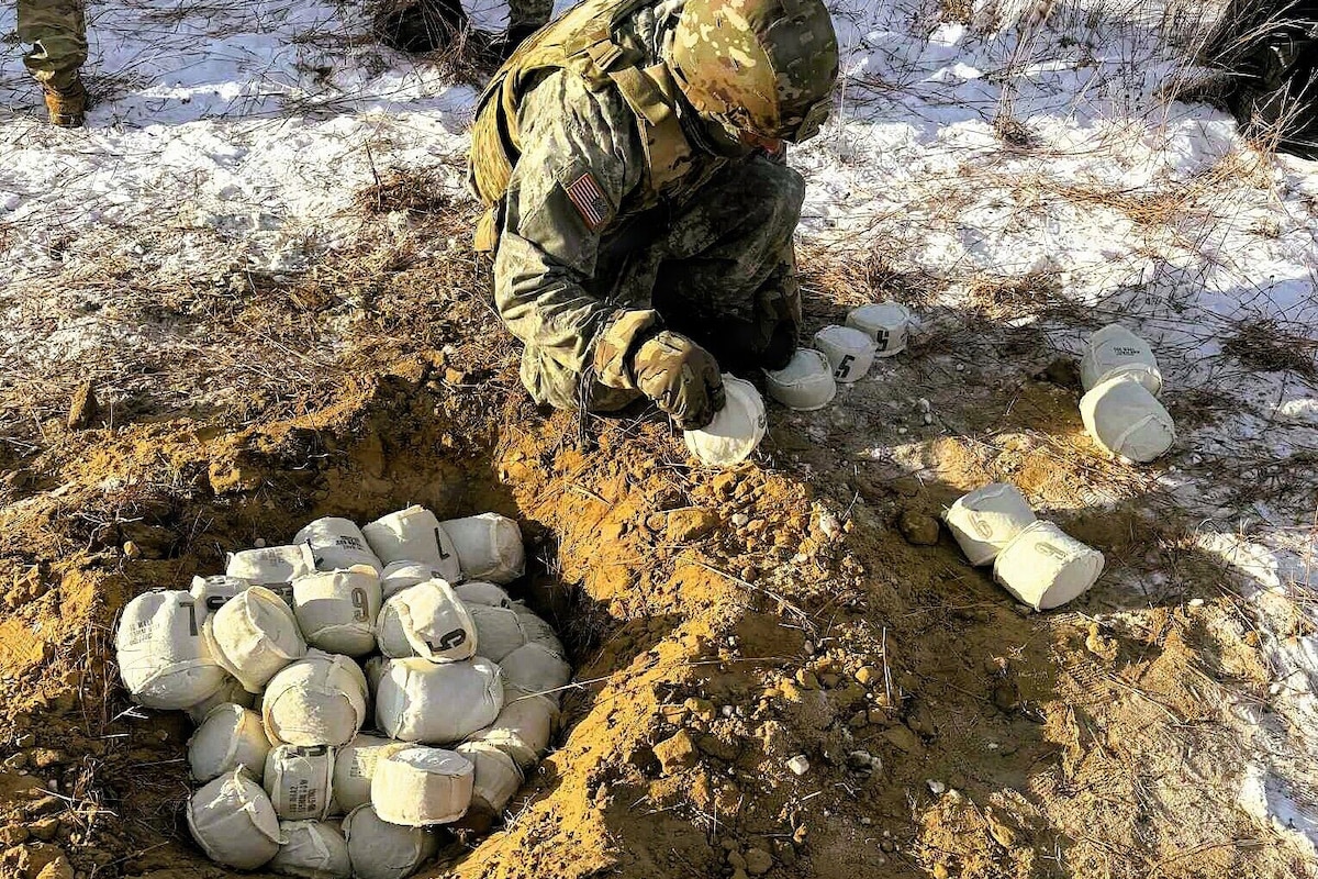 A man wearing a camouflage military uniform kneels in the snowy ground as he throws softball sized cloth pouches into a hole in the ground. There are several pouches in front of the man and a large pile in the hole.