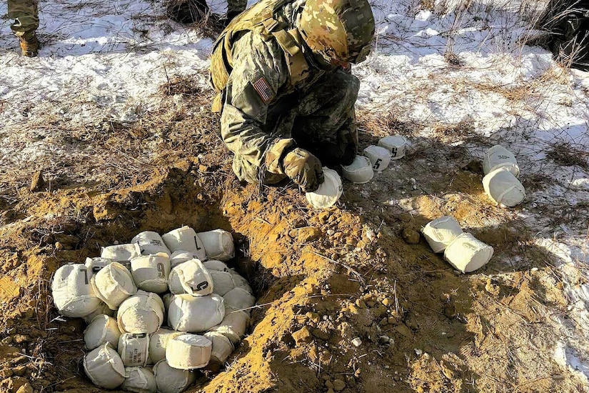 A man wearing a camouflage military uniform kneels in the snowy ground as he throws softball sized cloth pouches into a hole in the ground. There are several pouches in front of the man and a large pile in the hole.