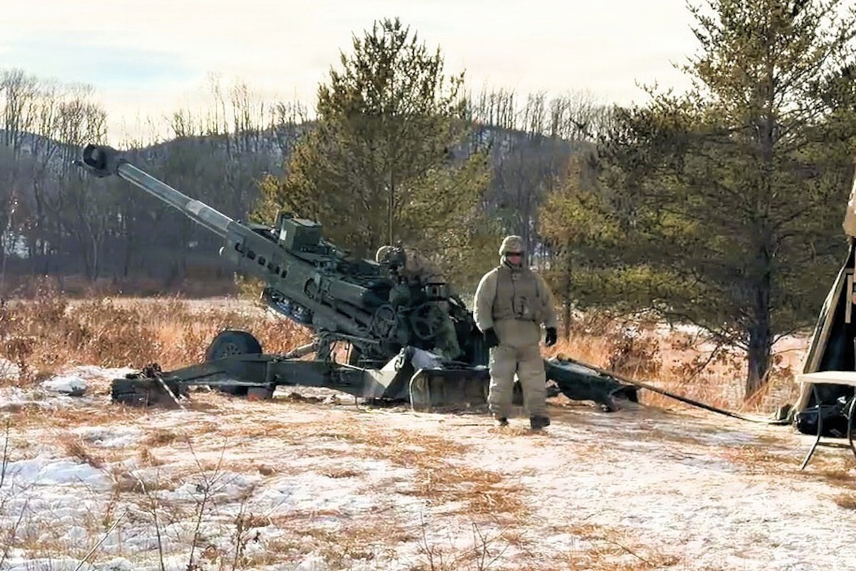 A person wearing a camouflage military uniform walks away from a large military artillery weapon stationed in the snow. There are trees and hills in the background.