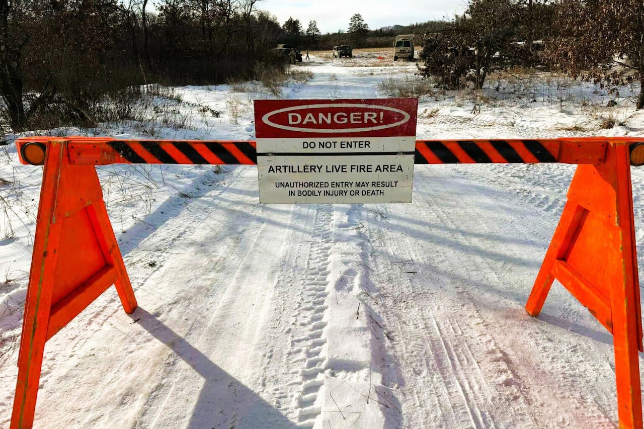 A barrier blocks a snow-covered road. A sign on the barrier reads, “Danger! Do not enter. Artillery live fire area.” There are three military vehicles parked at the end of the road.