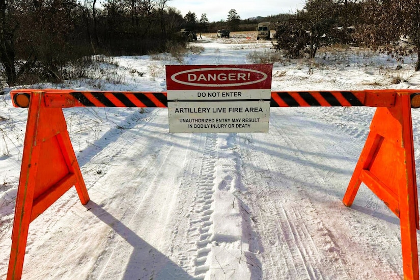 A barrier blocks a snow-covered road. A sign on the barrier reads, “Danger! Do not enter. Artillery live fire area.” There are three military vehicles parked at the end of the road.