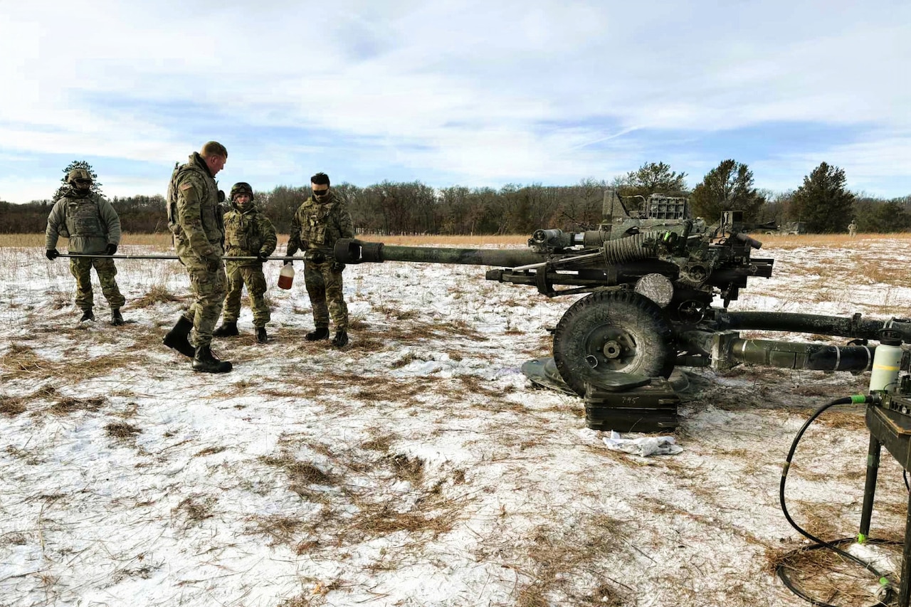 Four people wearing camouflage military uniforms load a large military artillery weapon stationed on a snowy field.