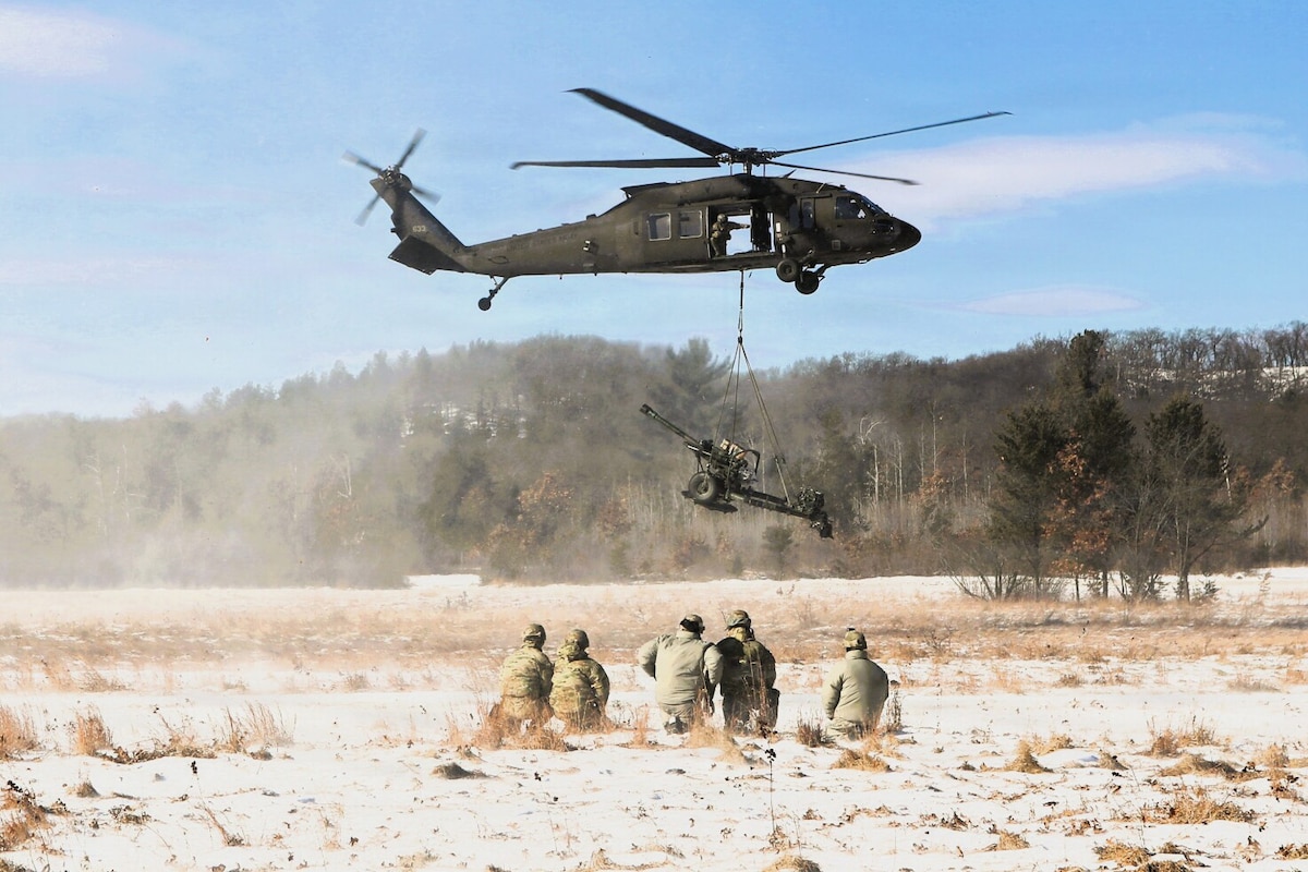 A military helicopter lowers a large artillery weapon to the ground on a snowy field. Five people wearing camouflage military uniforms are kneeling as they observe.
