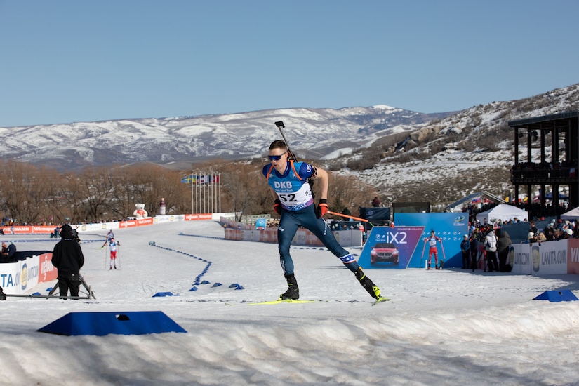 A man wearing sports attire skis along a flat, snowy course. Other skiers and spectators can be seen in the distance.