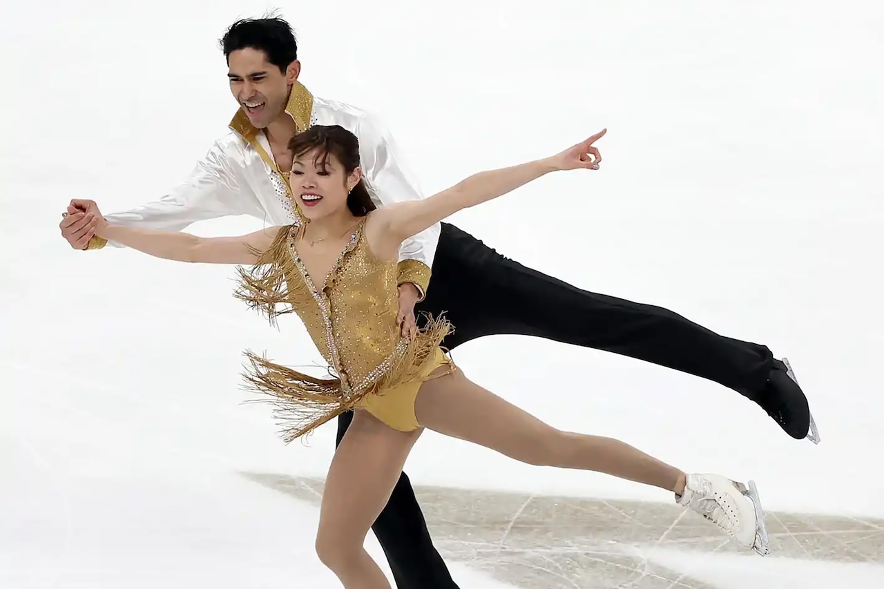 A man and a woman wearing ice skates and figure skating costumes hold hands and smile as they glide across ice, each on one leg with the other kicked behind them into the air.