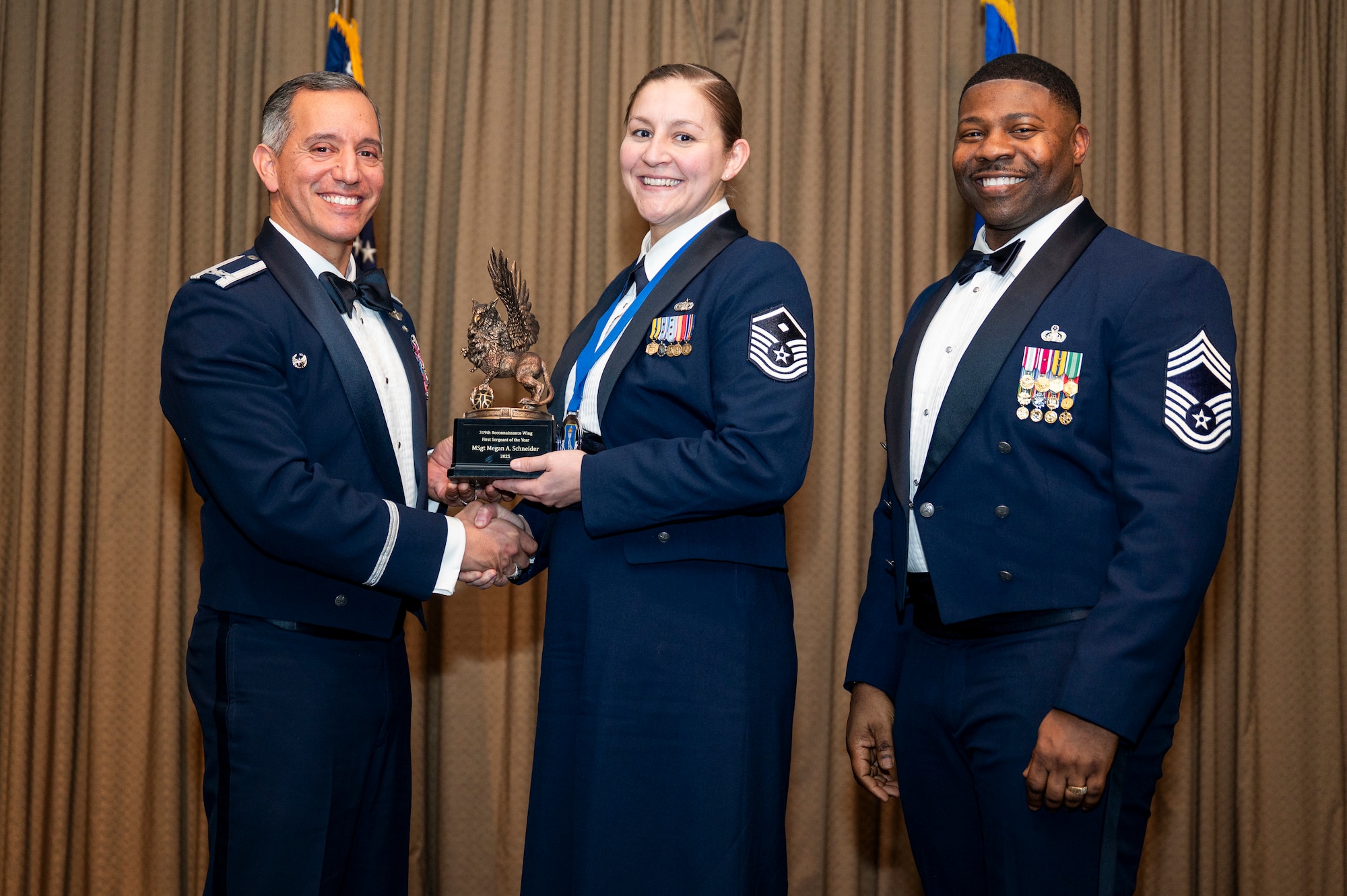 Col. Alfred Rosales, 319th Reconnaissance Wing commander, left and Chief Master Sgt. Marcus Cook Perry, 319th MSG command chief, right, present MSgt Megan A. Schneider, Wing Staff Agency, the First Sergeant of the year award during the 319th Reconnaissance Wing’s 2025 annual awards ceremony at Grand Forks Air Force Base, North Dakota, Jan. 29, 2026