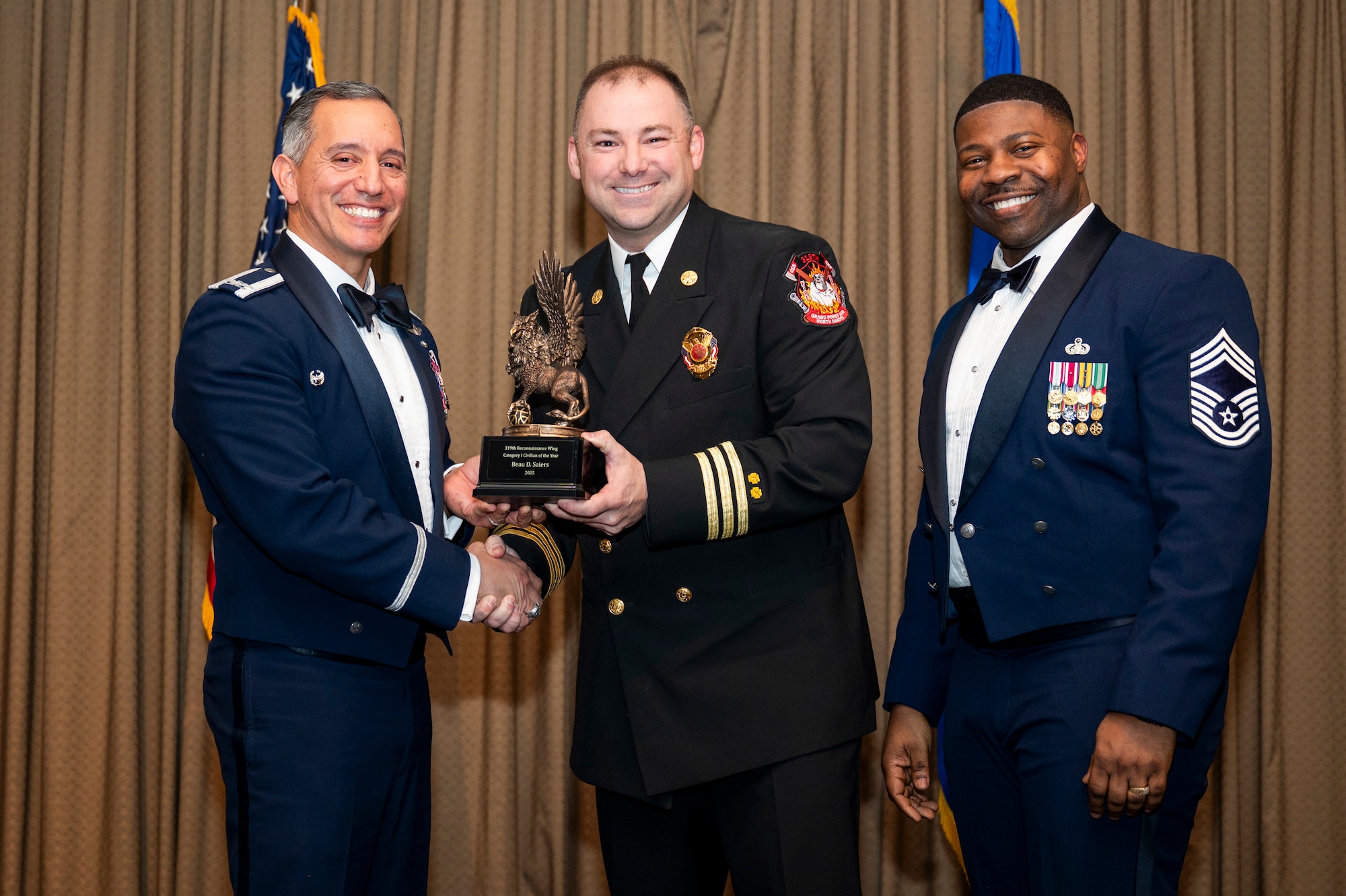 Col. Alfred Rosales, 319th Reconnaissance Wing commander, left and Chief Master Sgt. Marcus Cook Perry, 319th MSG command chief, right, present Beau D. Saiers, 319th Civil Engineer Squadron, the Category I Civilian of the Quarter Award, during the 319th Reconnaissance Wing’s 2025 annual awards ceremony at Grand Forks Air Force Base, North Dakota, Jan. 29, 2026.