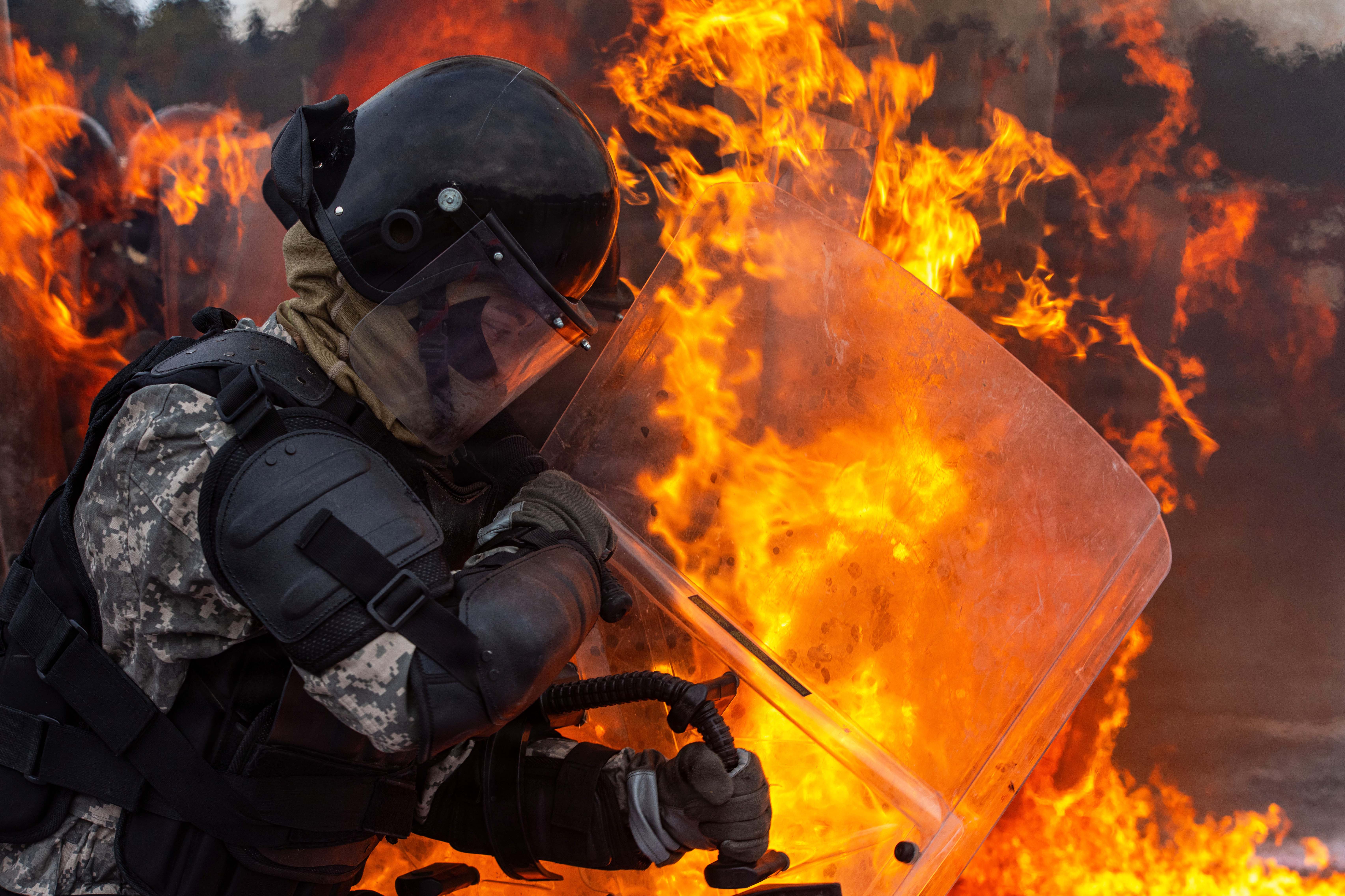 A soldier wearing a helmet and other protective gear holds a clear shield while navigating through flames.