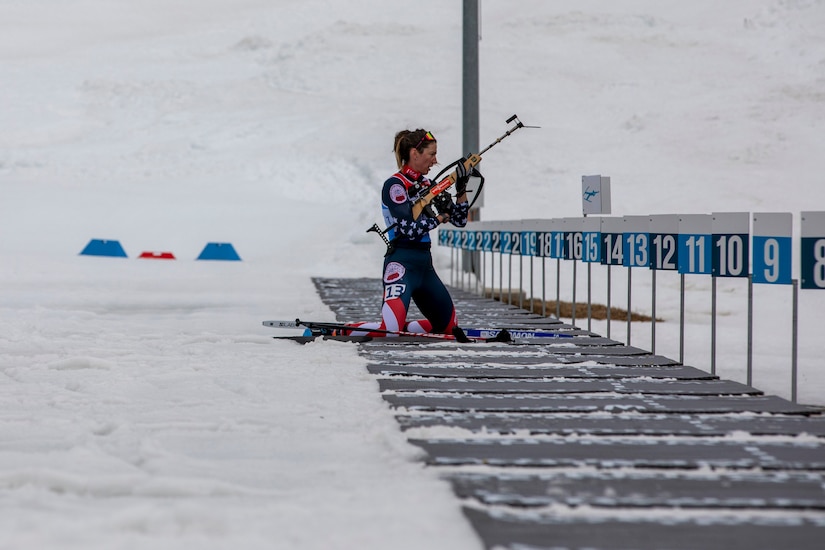 A woman wearing athletic attire and skis kneels in front of a line of numbered columns in the snow while preparing to aim a long gun.