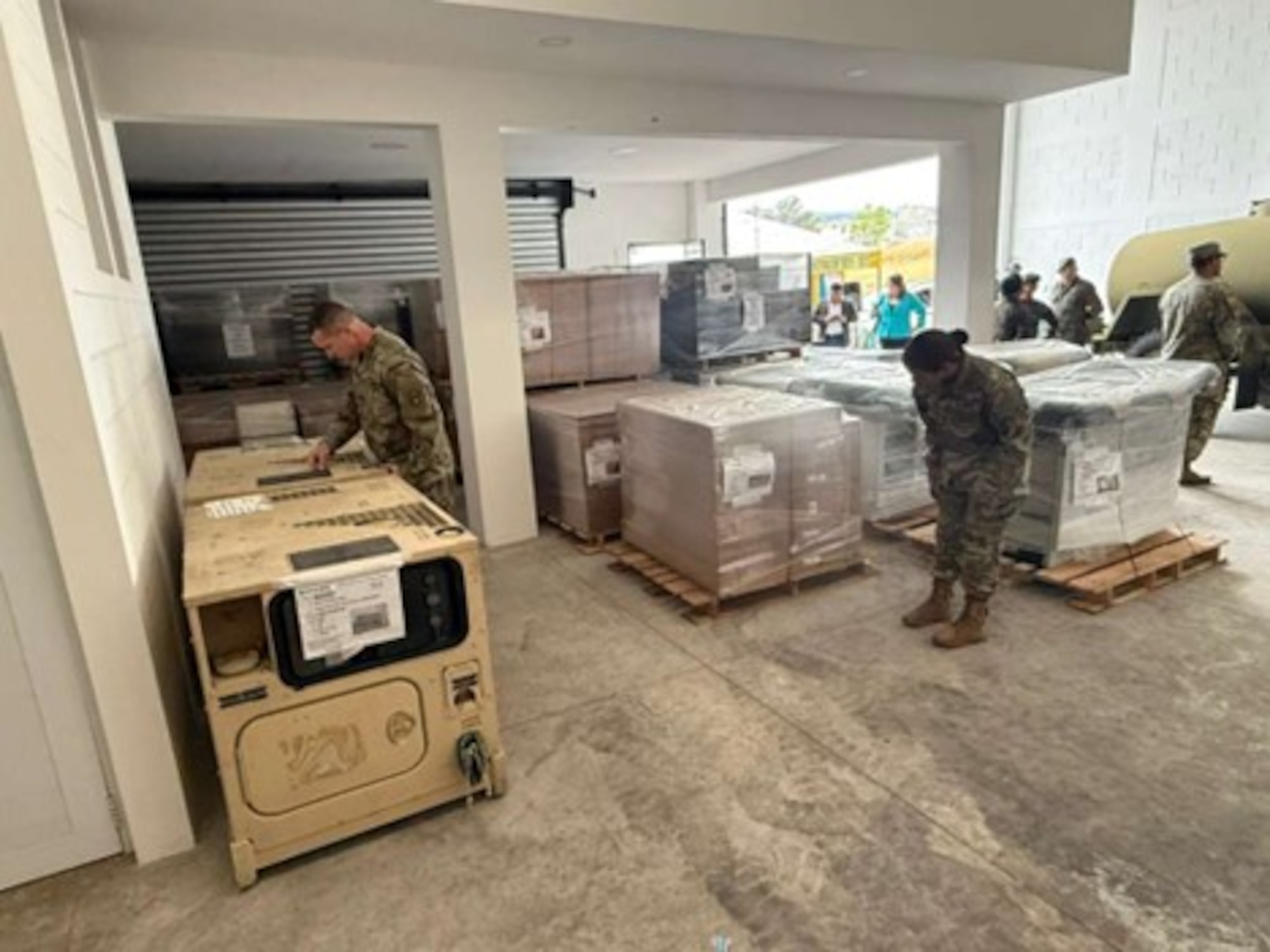 Military members stand in a warehouse with boxes and pallets.