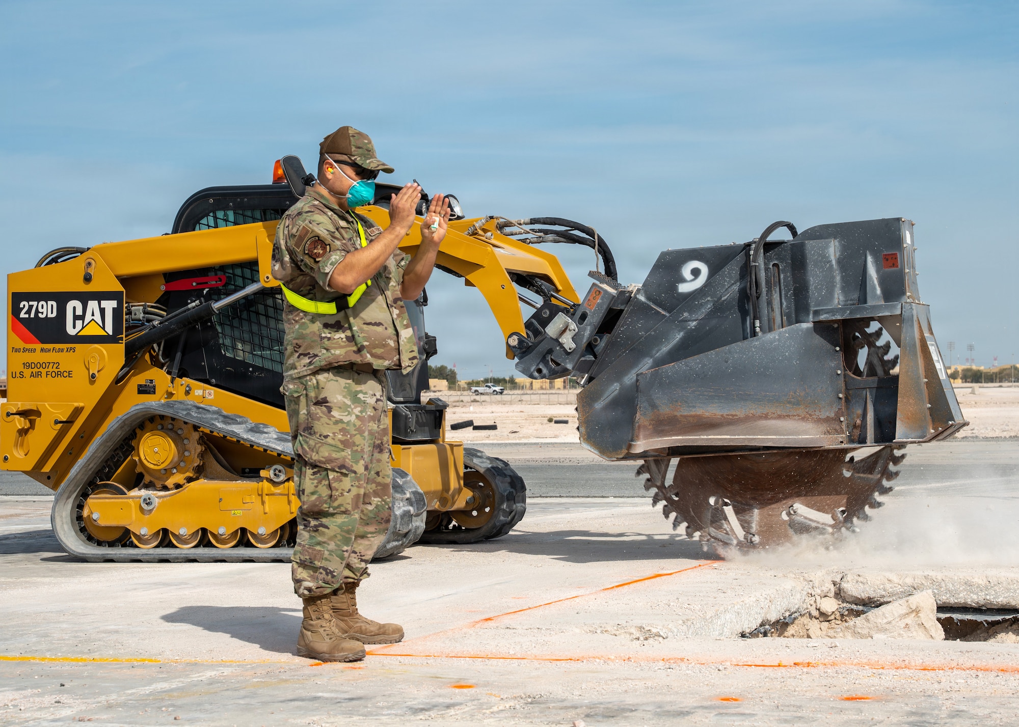 An Airman stands to the side of a hole in the ground while another operating heavy equipment with a large sawblade attached cuts into concrete