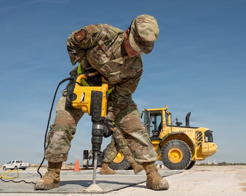 An Airman leans over a large drill and drills polymer matting to the ground. A bulldozer is parked in the background