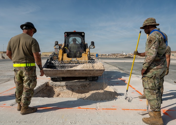 A bulldozer approaches a square pit with dirt while two Airmen stand next to the pit looking down