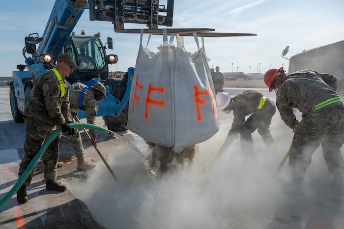 A forklift hoists a large container of cement mix over a pit creating a cloud of dust as it pours out. Airmen around the container rake the ground while one directs water from a hose into the pit