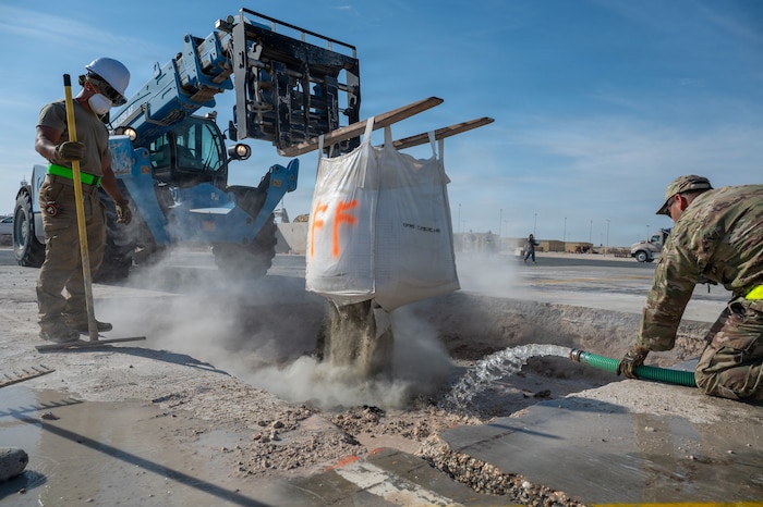 A large forklift hoists a bag of concrete mix over a hole while the mix pours into the hole. One Airman stands on the edge of the pit holding a rake while another kneels and directs water from a hose into the pit