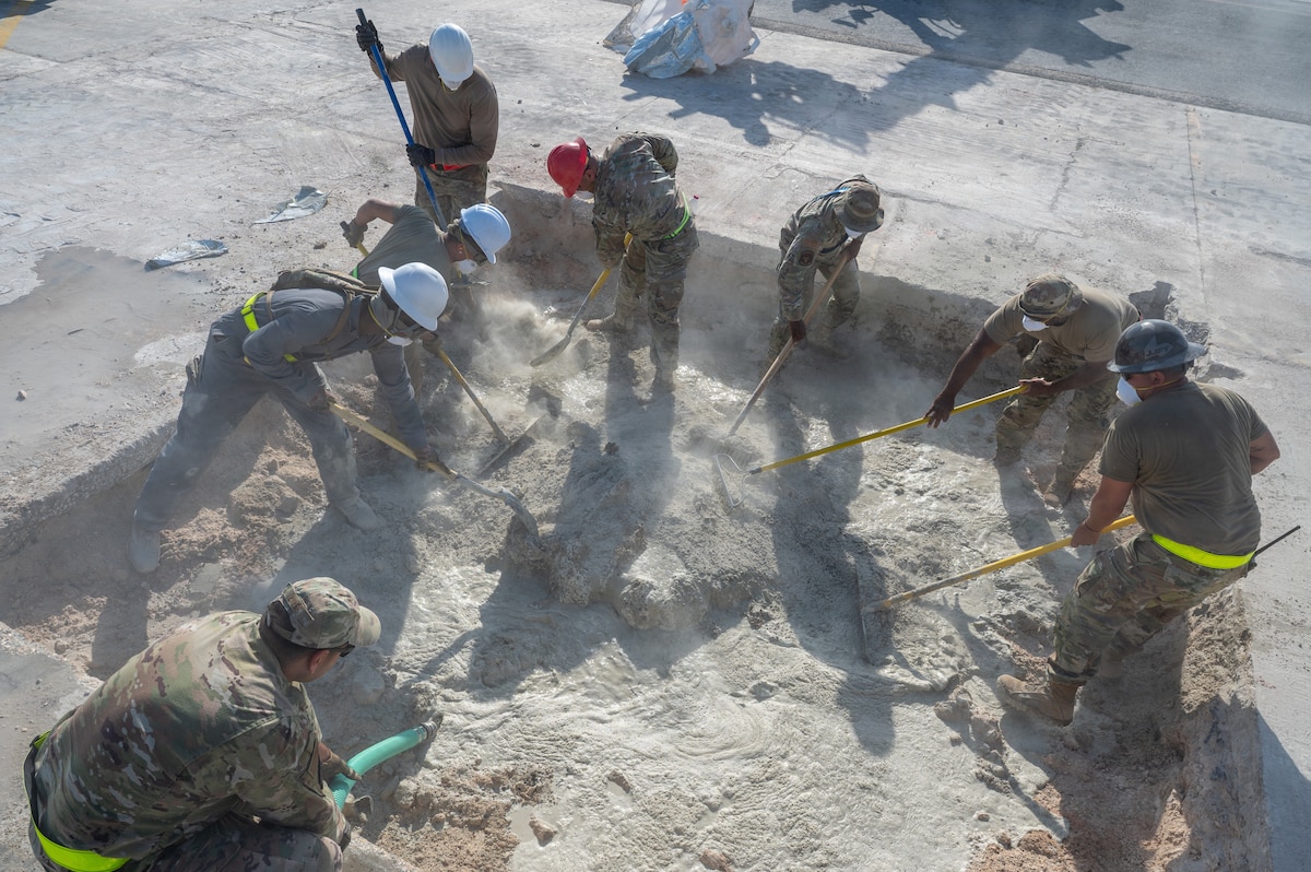 6 Airmen stand in a square shaped pit raking concrete while one Airman is on the edge of the square holding a water hose dispensing water into the pit