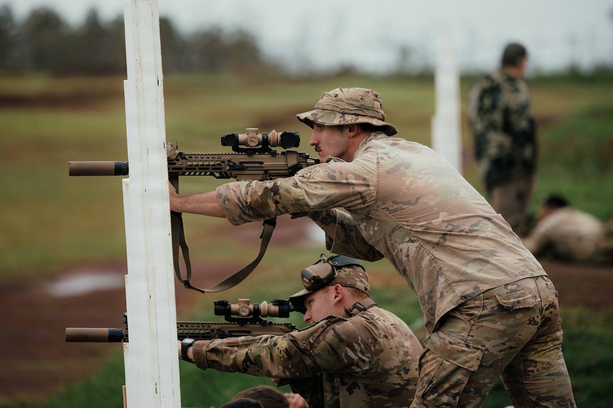 Two men in camouflage military uniforms, one standing and one kneeling in a grassy terrain, aim military rifles out of frame; the rifles are balanced on white support structures. Three other people in similar attire are doing the same in the background.