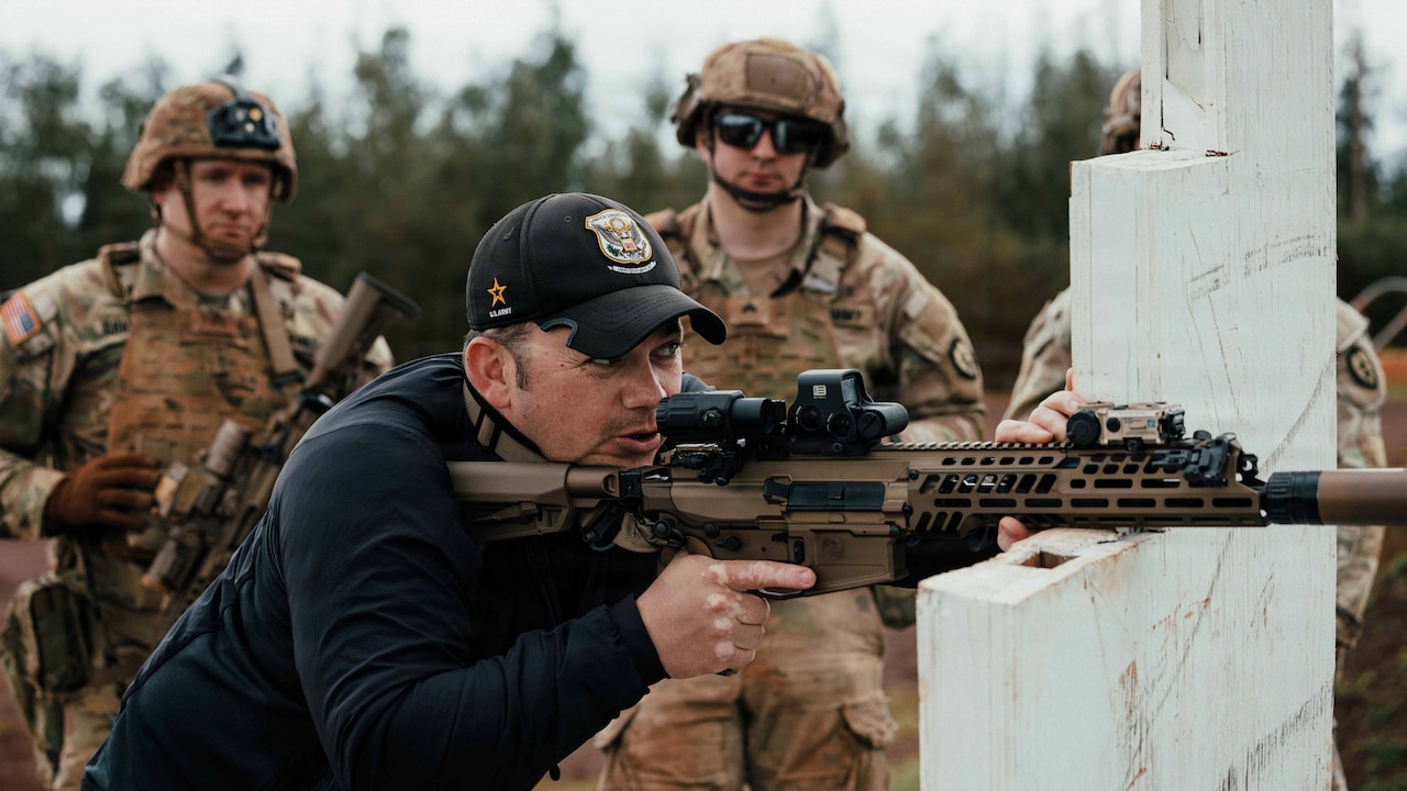 Three people in camouflage military uniforms and helmets stand and observe a man wearing casual attire and a baseball cap as he stands and aims a military rifle out of frame; the rifle is balanced on a white support structure, and trees are in the background.