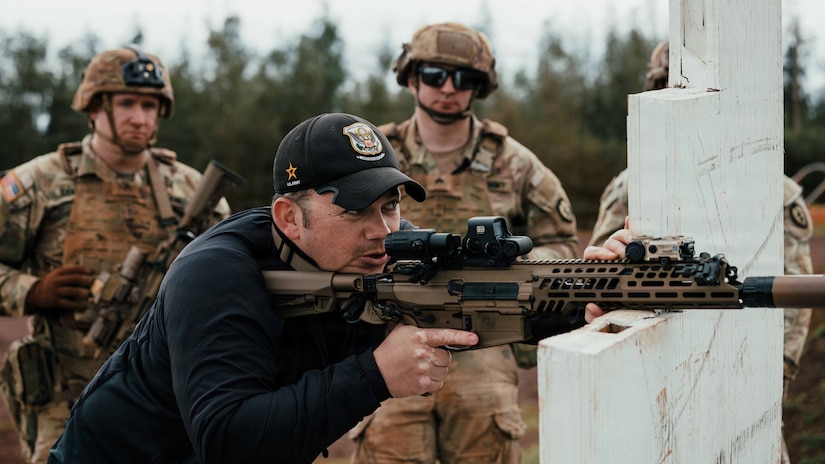 Three people in camouflage military uniforms and helmets stand and observe a man wearing casual attire and a baseball cap as he stands and aims a military rifle out of frame; the rifle is balanced on a white support structure, and trees are in the background.