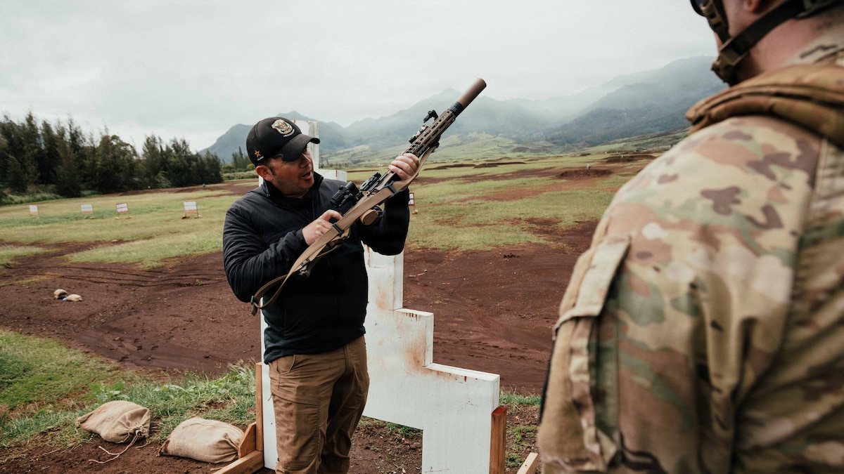 A man in casual attire and a baseball cap holds a military rifle outside in grassy terrain on a cloudy day with trees in the background, as a person in a camouflage military uniform stands partially out of frame and observes.