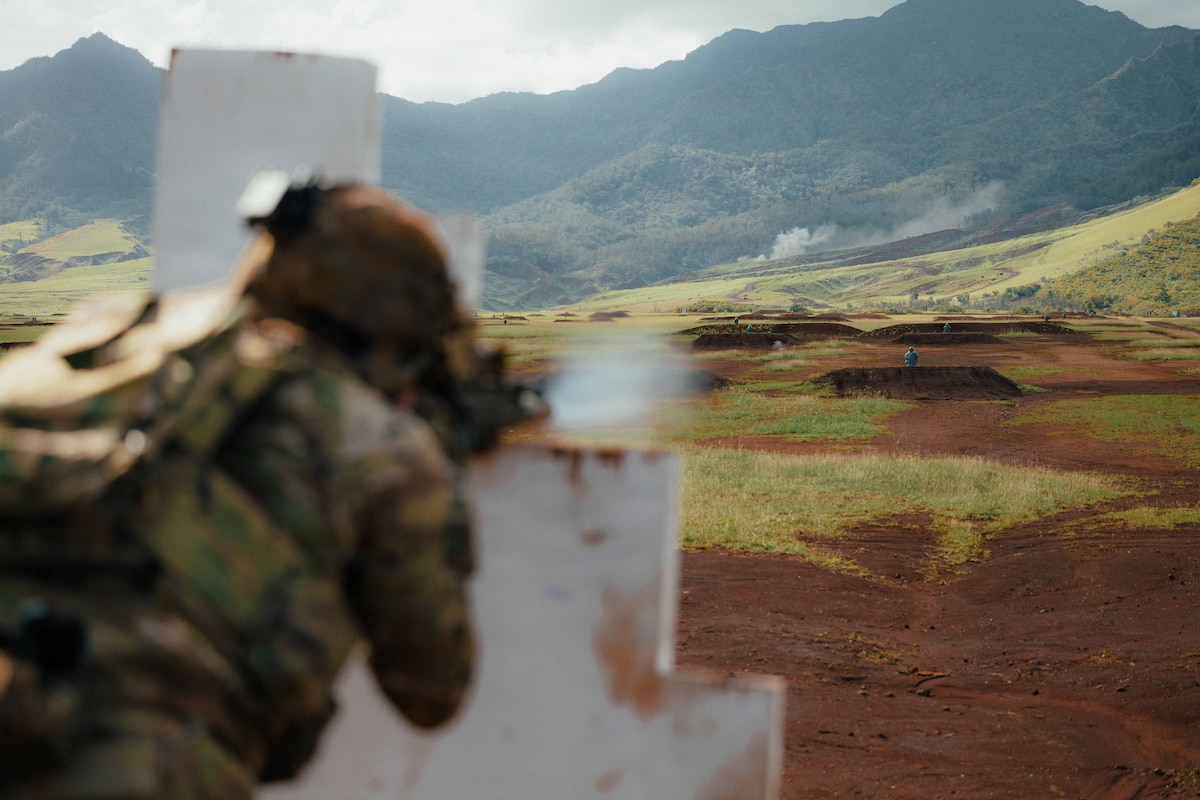 Two men in camouflage military uniforms, one standing and one kneeling in a grassy terrain, aim military rifles out of frame; the rifles are balanced on white support structures. Three other people in similar attire are doing the same in the background.