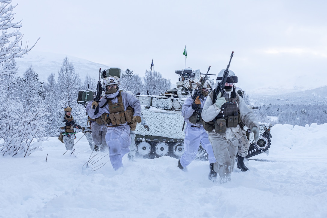 Marines and other service members in cold-weather tactical gear and carrying rifles move through snow, with a tank, trees and mountains in the background.