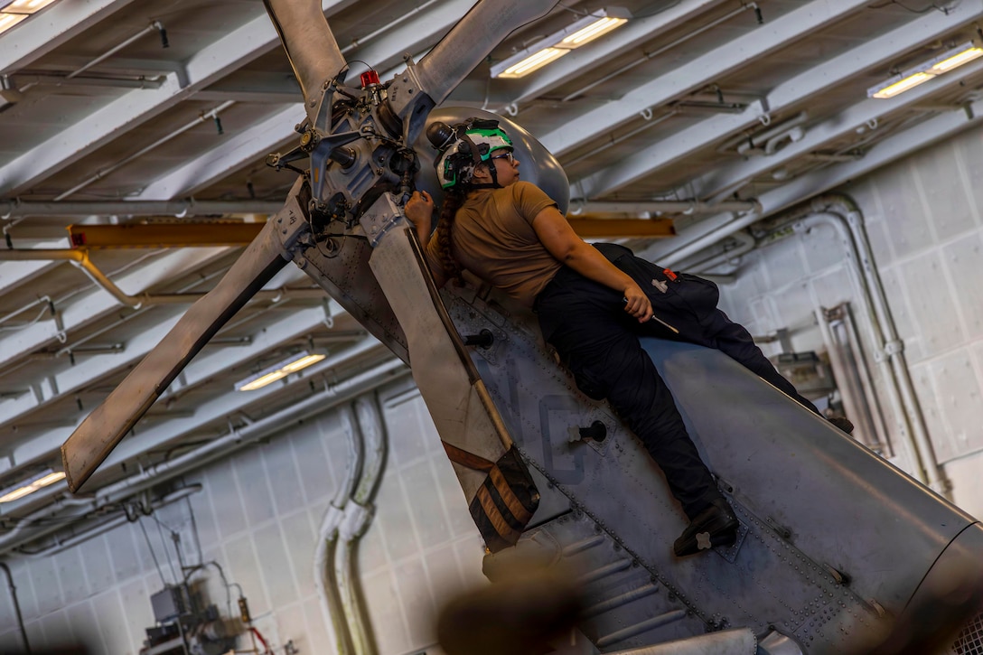 A person wearing civilian attire and a helmet looks to the right while holding a tool and straddling a helicopter near the rear propeller.