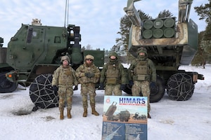 Four people wearing camouflage military uniforms stand shoulder to shoulder while posing for a photo in from of a rocket launching system.