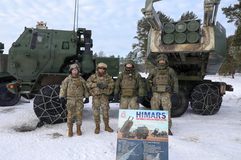 Four people wearing camouflage military uniforms stand shoulder to shoulder while posing for a photo in from of a rocket launching system.