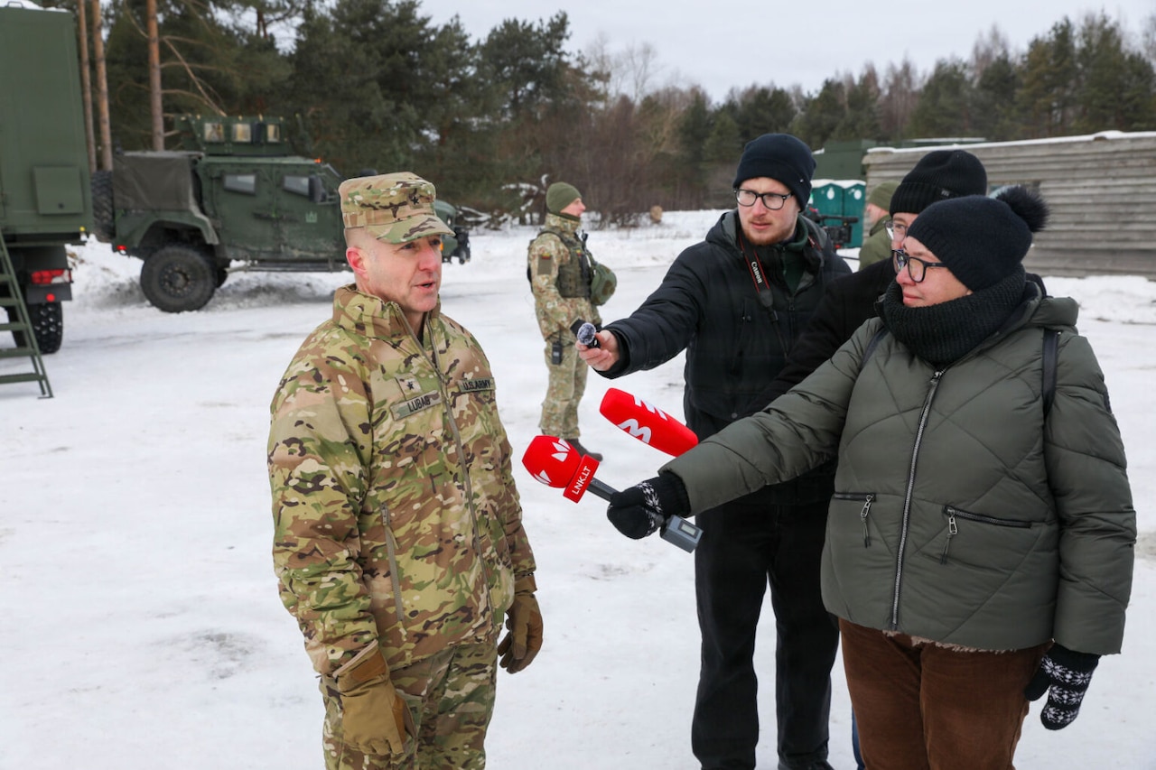 A woman and two men wearing winter apparel hold microphones in front of a man dressed in a camouflage military uniform as they all stand in a snow-covered field. There is another man in a camouflage military uniform standing in the background with military vehicles and equipment behind him.