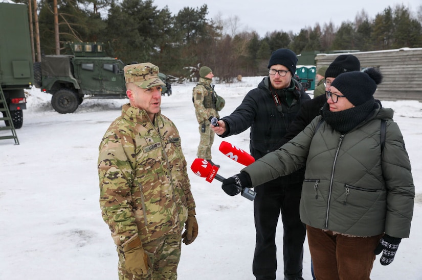 A woman and two men wearing winter apparel hold microphones in front of a man dressed in a camouflage military uniform as they all stand in a snow-covered field. There is another man in a camouflage military uniform standing in the background with military vehicles and equipment behind him.