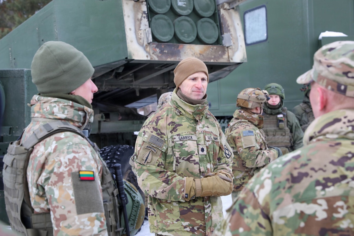A man wearing a camouflage military uniform speaks to several others dressed in camouflage military uniforms. There is a rocket launching system in the background.