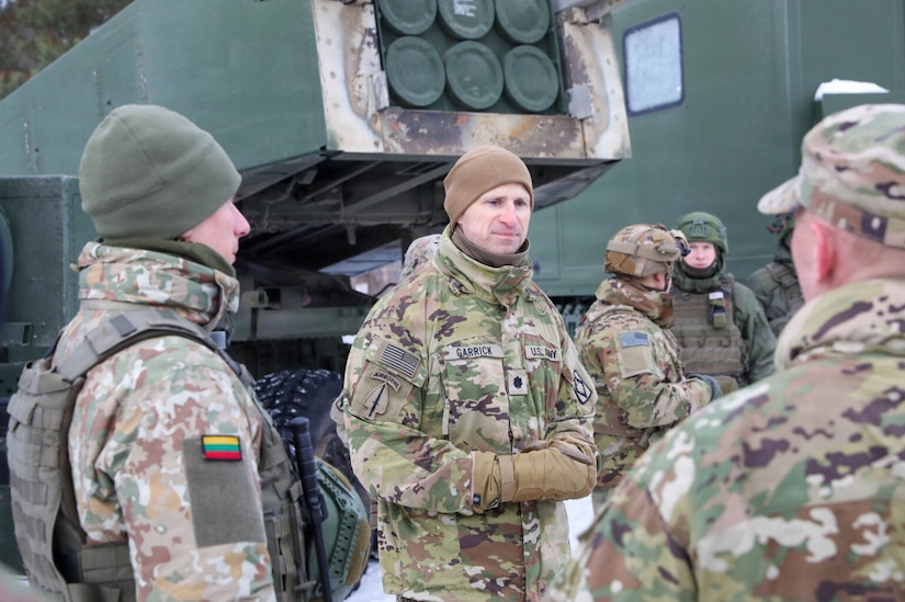 A man wearing a camouflage military uniform speaks to several others dressed in camouflage military uniforms. There is a rocket launching system in the background.