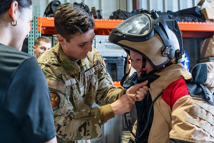 An Airman helps a student put on a helmet on a bomb suit