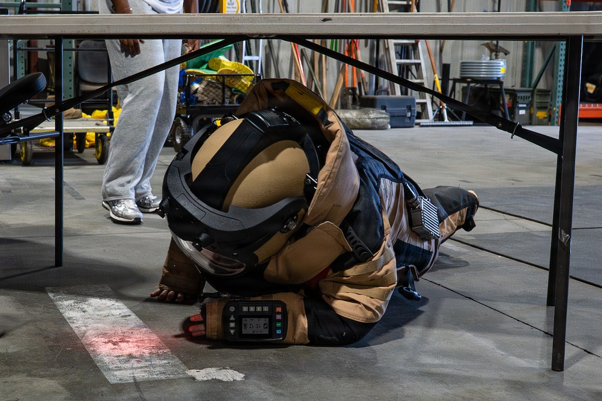 A student wearing a bomb suit crawls under a table