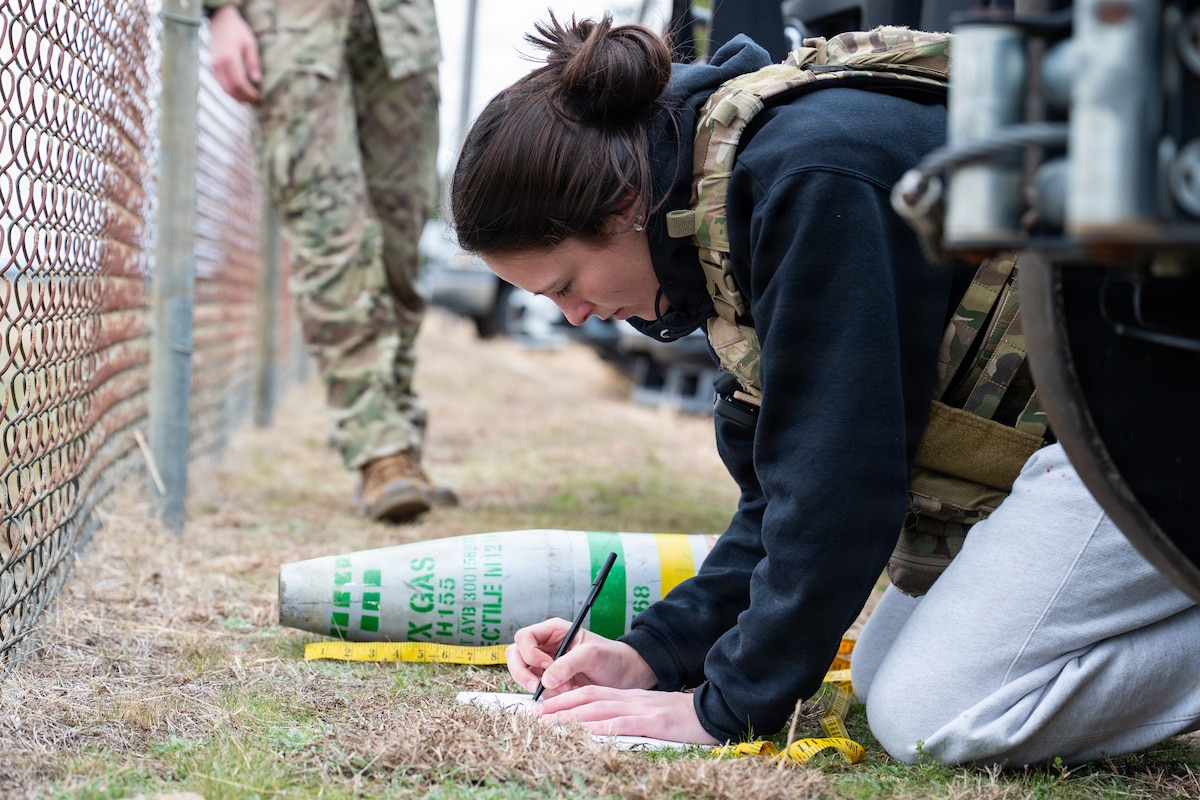 A student wearing a flak vest kneels downs and writes notes on paper by a ruler and a mock bomb