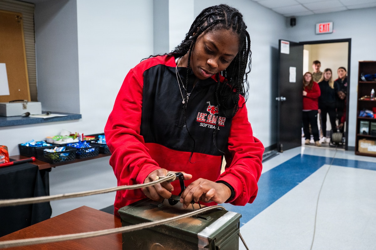 A student attaches a carabiner tied to a rope to an ammo can on a table